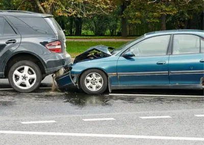 A blue sedan has rear-ended a black SUV on a wet road, causing visible front-end damage to the sedan and rear damage to the SUV. Trees and greenery are in the background.