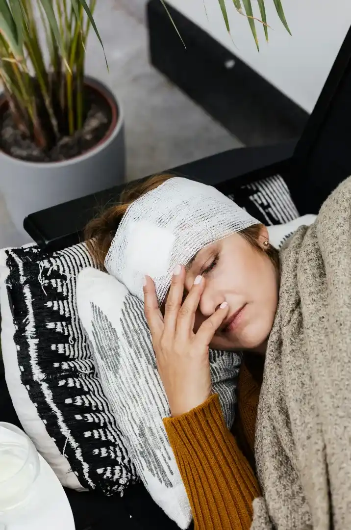 A person with a bandaged head lies on a pillow, covered with a blanket, resting their hand on their forehead.